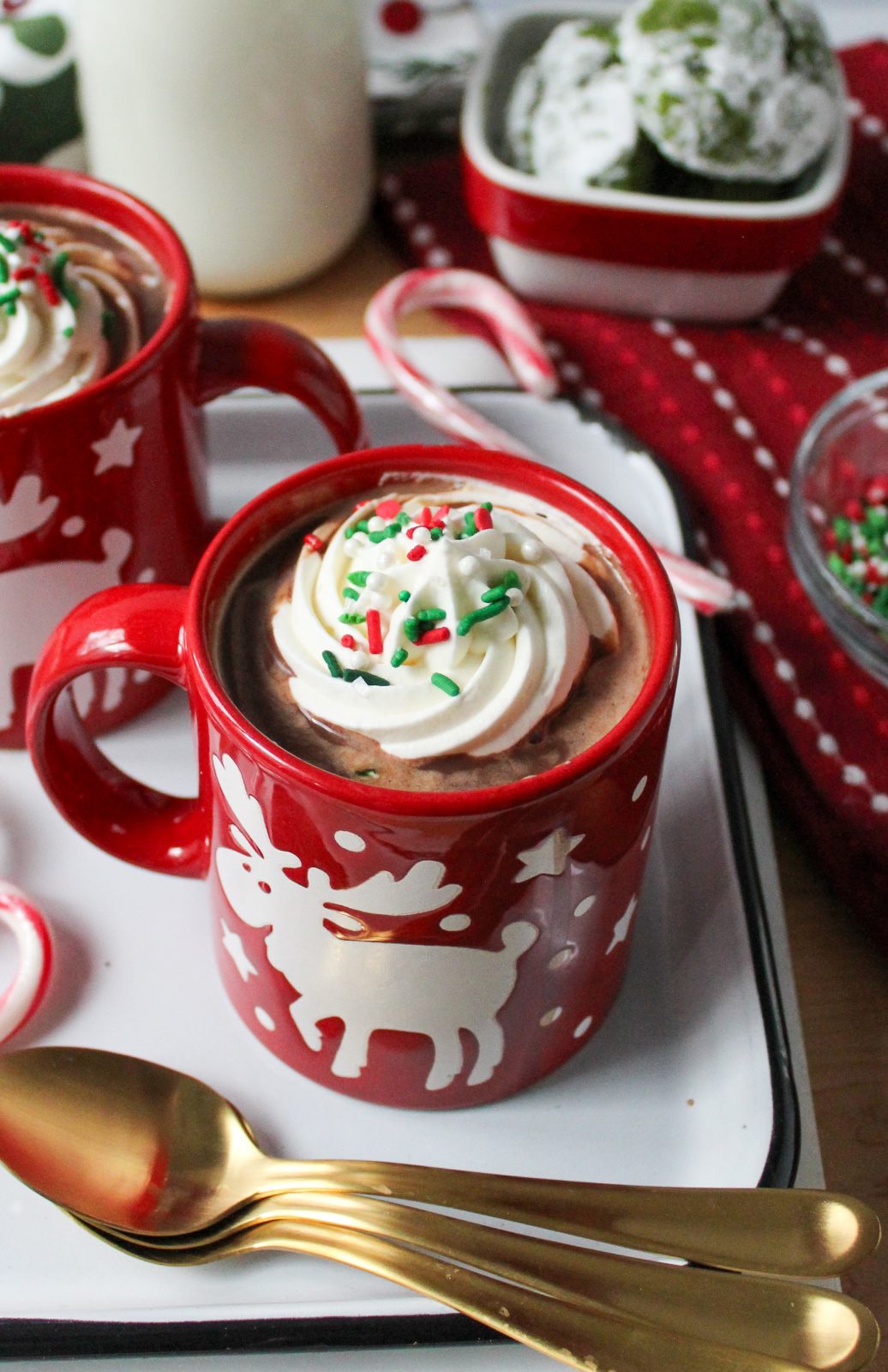 Recipe Photo for Frozen Marshmallow Whipped Cream Cocoa Toppers. Portrait orientation photo shows red moose mugs with cocoa and the frozen toppers floating on the cocoa. The mugs are on a vintage enamel tray with gold spoons and candy canes. In the background is a small bowl with green crinkle cookies and a small jar with milk.