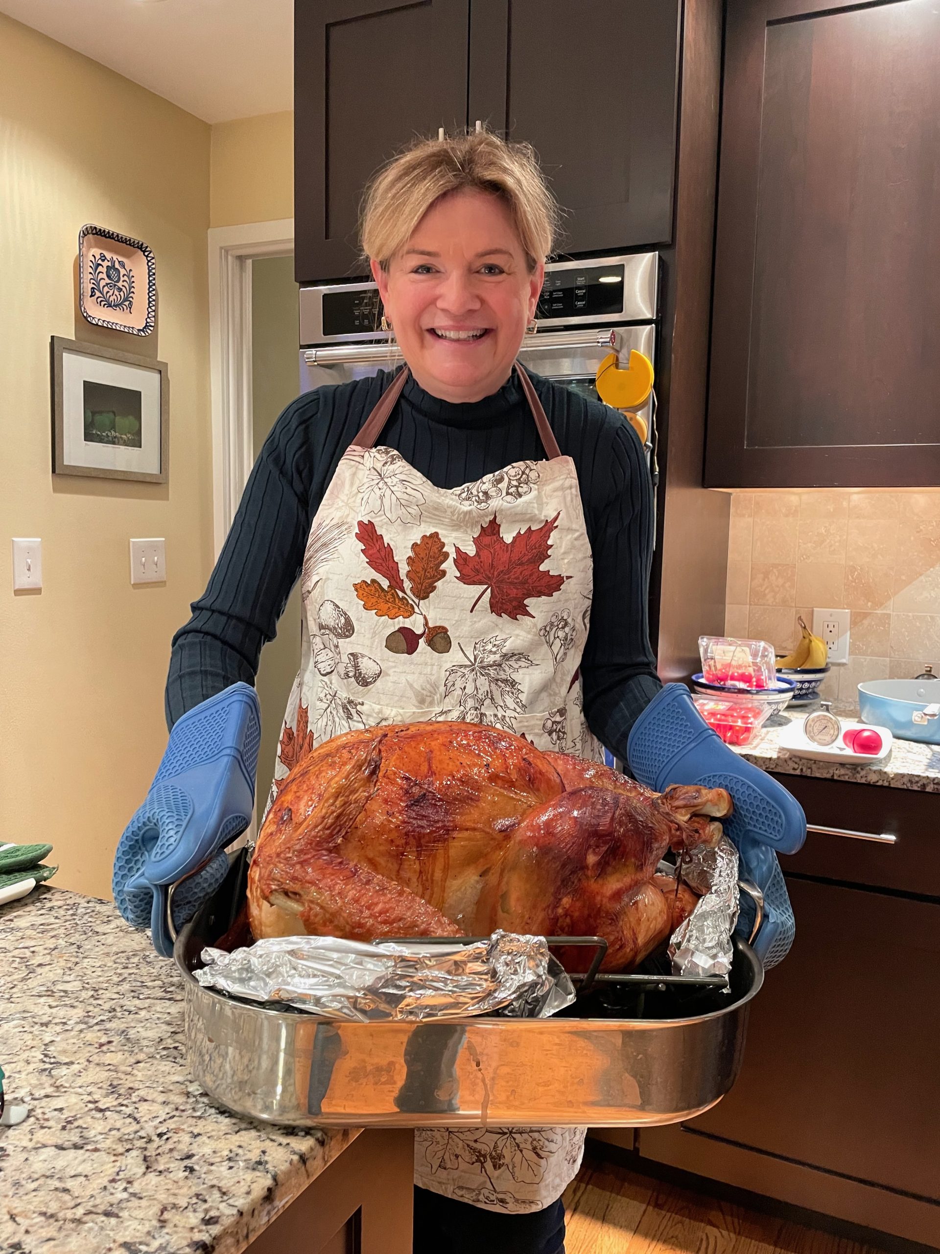 Photo of Tina Verrelli - food blogger and cookbook author epicuricloud.com holding a roasted Thanksgiving turkey in her kitchen. She's wearing a fall patterned apron with autumn leaves.