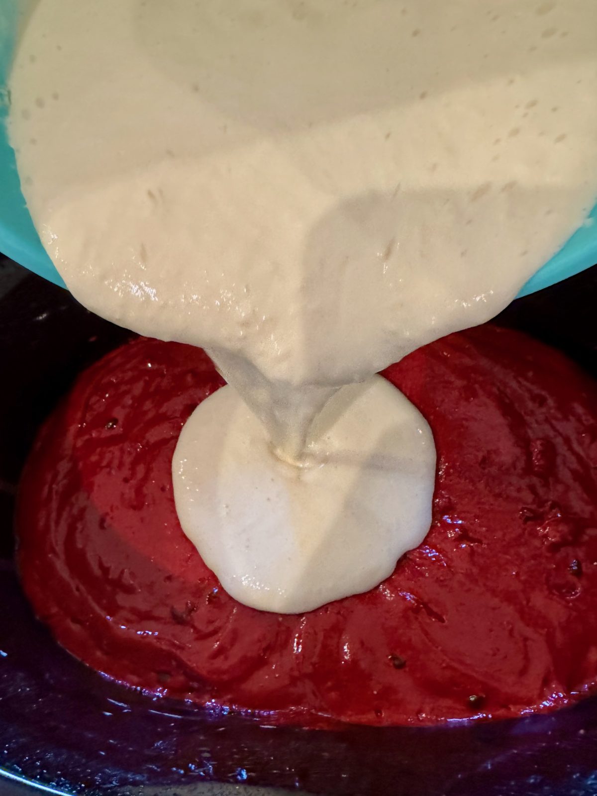 Recipe Photo for Red Velvet White Chocolate (Slow Cooker) Lava Cake. A ice blue KitchenAid Mixing bowl containing the cream cheese mixture is being poured over the mixed red velvet cake mixture in the black slow cooker.