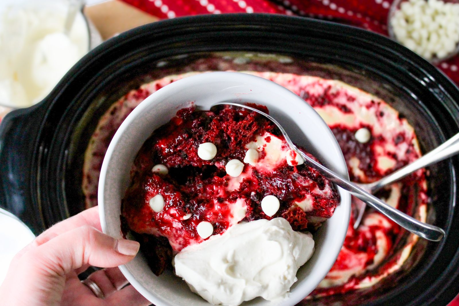 Recipe Photo for Red Velvet White Chocolate (Slow Cooker) Lava Cake.  Portrait orientation photo showing a hand holding a white bowl of Lava cake with whipped cream and a sprinkling of white chocolate chips. held over a black slow cooker with more of the red and white colored cake mixture.  A red and white dot patterned cloth napkin is in the background.