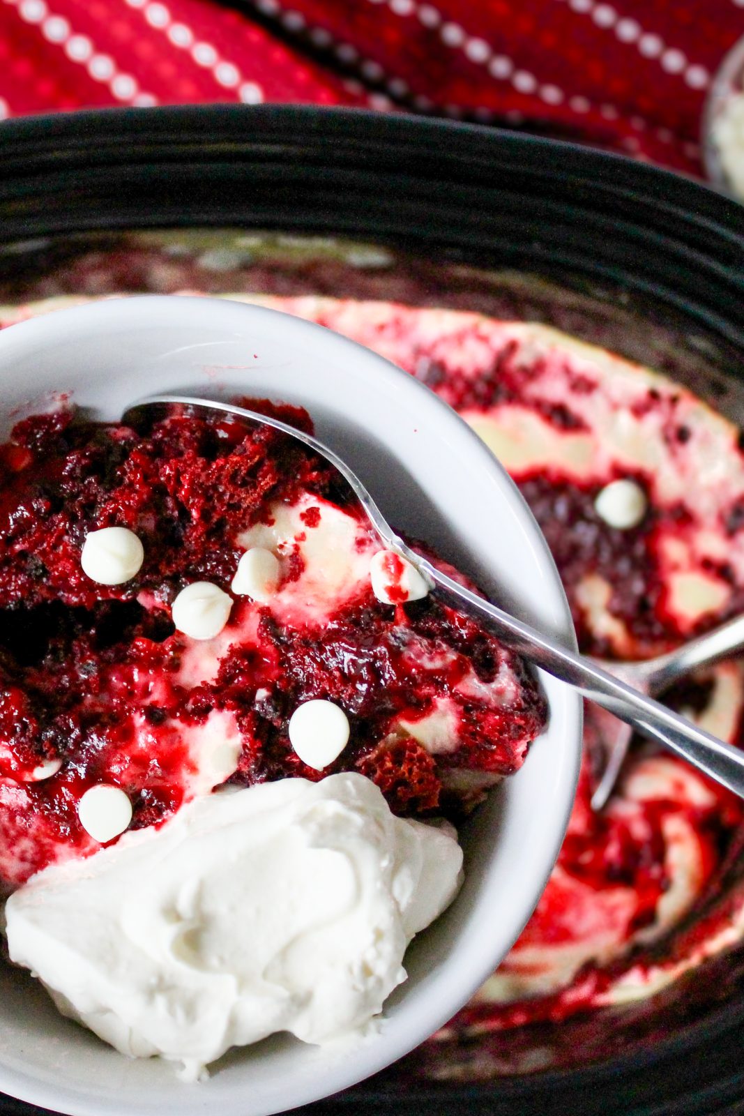 Recipe Photo for Red Velvet White Chocolate Slow Cooker Lava Cake. Portrait orientation photo showing white bowl of Lava cake with whipped cream and a sprinkling of white chocolate chips. held over a black slow cooker with more of the red and white colored cake mixture. A red and white dot patterned cloth napkin is in the background.
