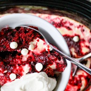 Recipe Photo for Red Velvet White Chocolate Slow Cooker Lava Cake. Portrait orientation photo showing white bowl of Lava cake with whipped cream and a sprinkling of white chocolate chips. held over a black slow cooker with more of the red and white colored cake mixture. A red and white dot patterned cloth napkin is in the background.