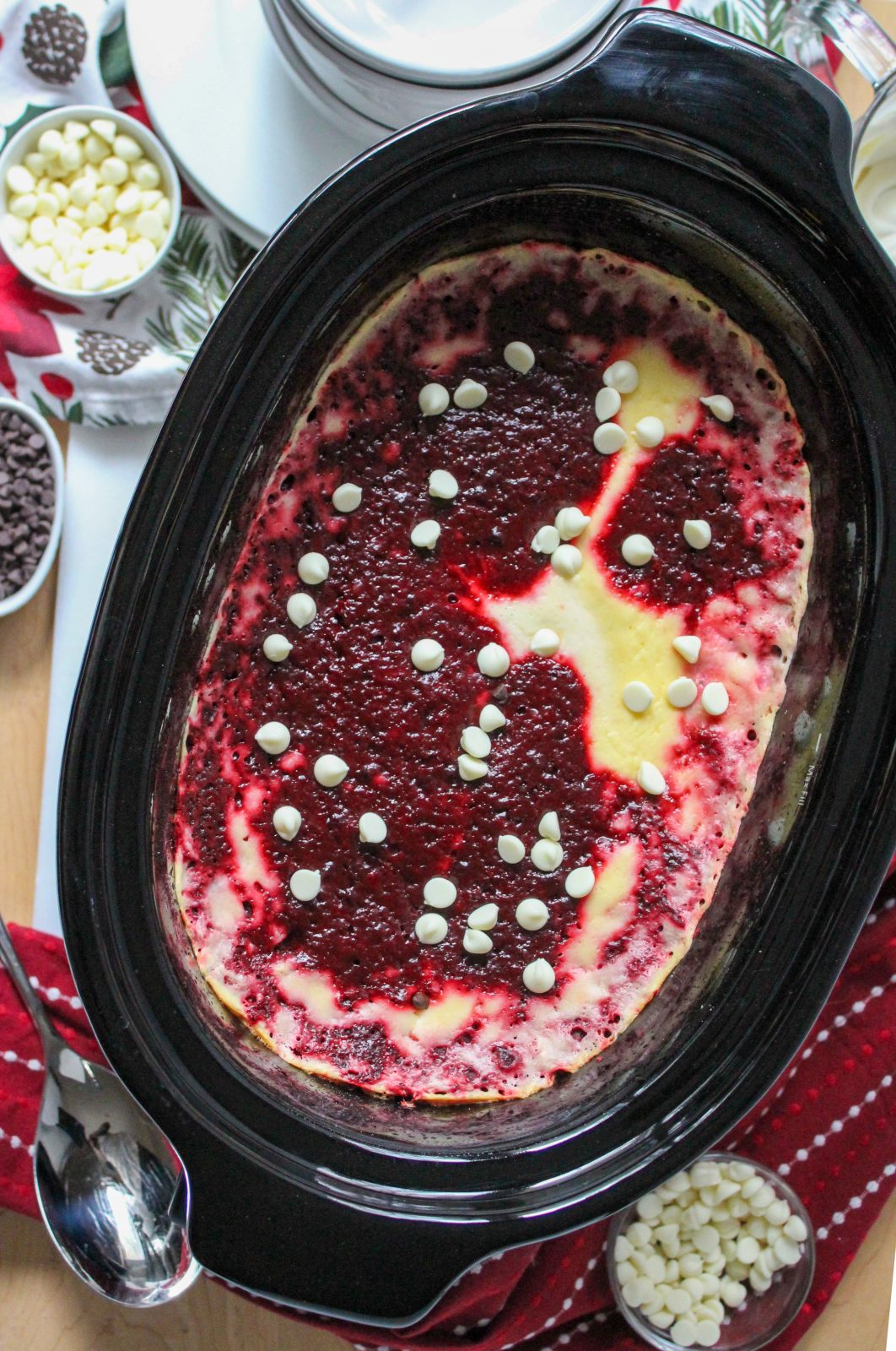 Recipe Photo for Red Velvet White Chocolate (Slow Cooker) Lava Cake.  Portrait orientation photo showing black slow cooker with the baked red and white swirled cake mixture with a sprinkle of white chocolate chips overtop.  A red and white dot patterned cloth napkin is in the background along with a bowl of whipped cream and a small dish filled with white chocolate chips.