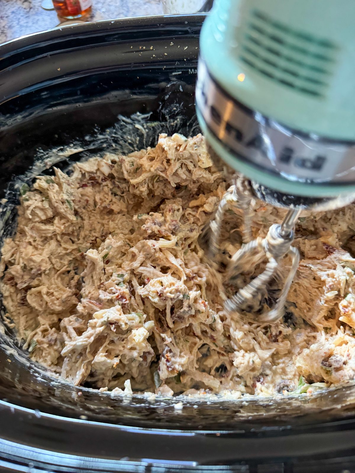 Recipe Photo for Marry Me Chicken Dip Showing all the ingredients for the dip in a black oval slow cooker. A KitchenAid Hand Mixer is just finished mixing them: Cream cheese cubes, chopped sun dried tomatoes, Parmesan and mozzarella cheeses, ranch dressing, fresh basil and seasonings.