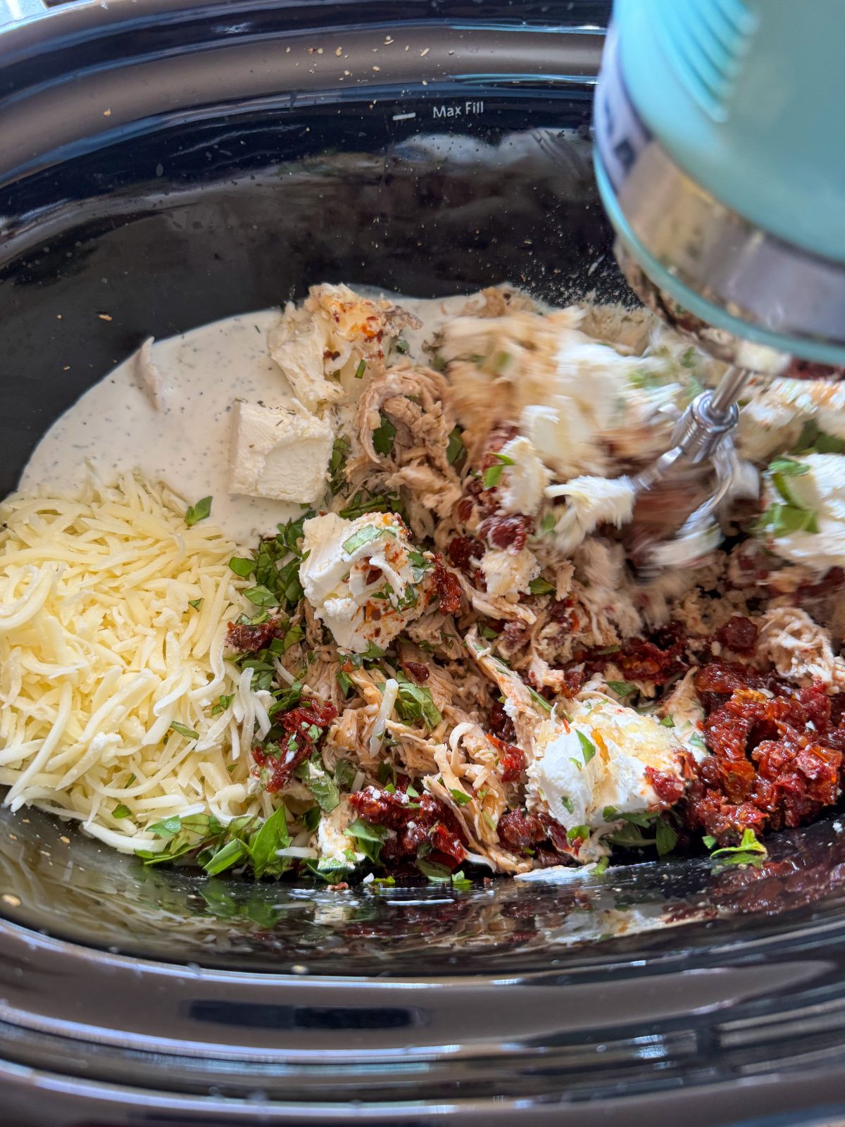 Recipe Photo for Marry Me Chicken Dip Showing all the ingredients for the dip in a black oval slow cooker. A KitchenAid Hand Mixer is starting to mix them: Cream cheese cubes, chopped sun dried tomatoes, Parmesan and mozzarella cheeses, ranch dressing, fresh basil and seasonings.