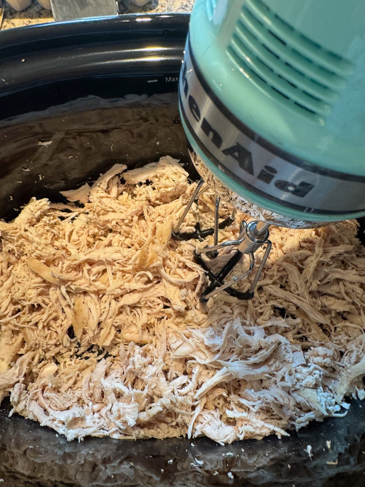 Recipe Photo for Marry Me Chicken Dip Showing a KitchenAid Hand Mixer almost done shredding chicken breasts in a black oval slow cooker.