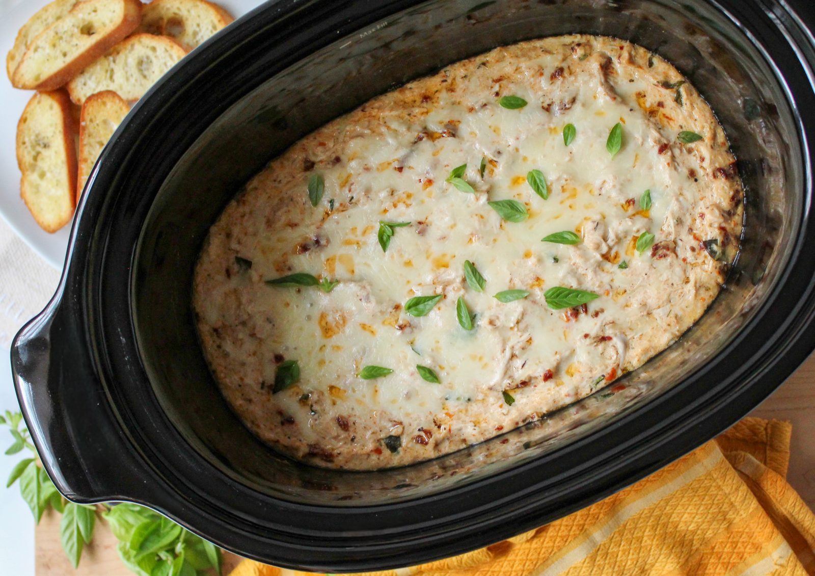 Recipe photo (landscape orientation) for Marry Me Chicken Dip. Showing final baked dip in an oval black slow cooker stoneware insert, surrounded by a gold kitchen towel, fresh basil sprigs and toasted crostini. The dip is cheese topped and baked golden brown.