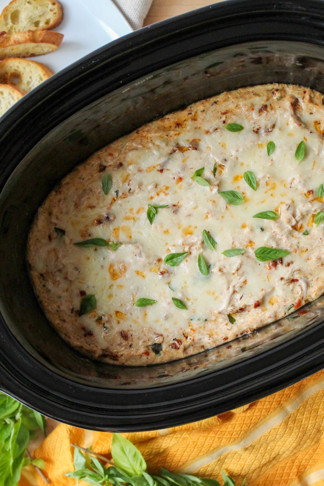 Recipe photo (portrait orientation) for Marry Me Chicken Dip. Showing final baked dip in an oval black slow cooker stoneware insert, surrounded by a gold kitchen towel, fresh basil sprigs and toasted crostini. The dip is cheese topped and baked golden brown.