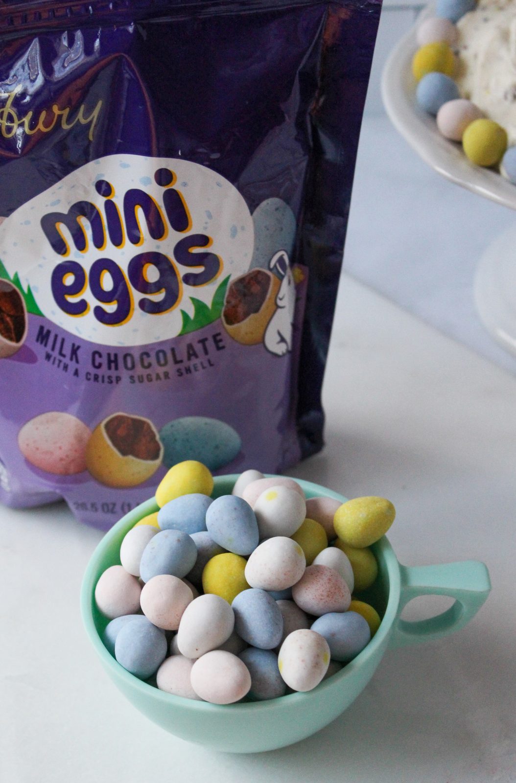 Image showing a purple package for Cadbury Mini Eggs. A vintage plastic aqua colored cup filled with Cadbury Mini Eggs, in the background you can glimpse Cadbury Mini Egg Whipped Cheesecake Dip in a footed bowl.