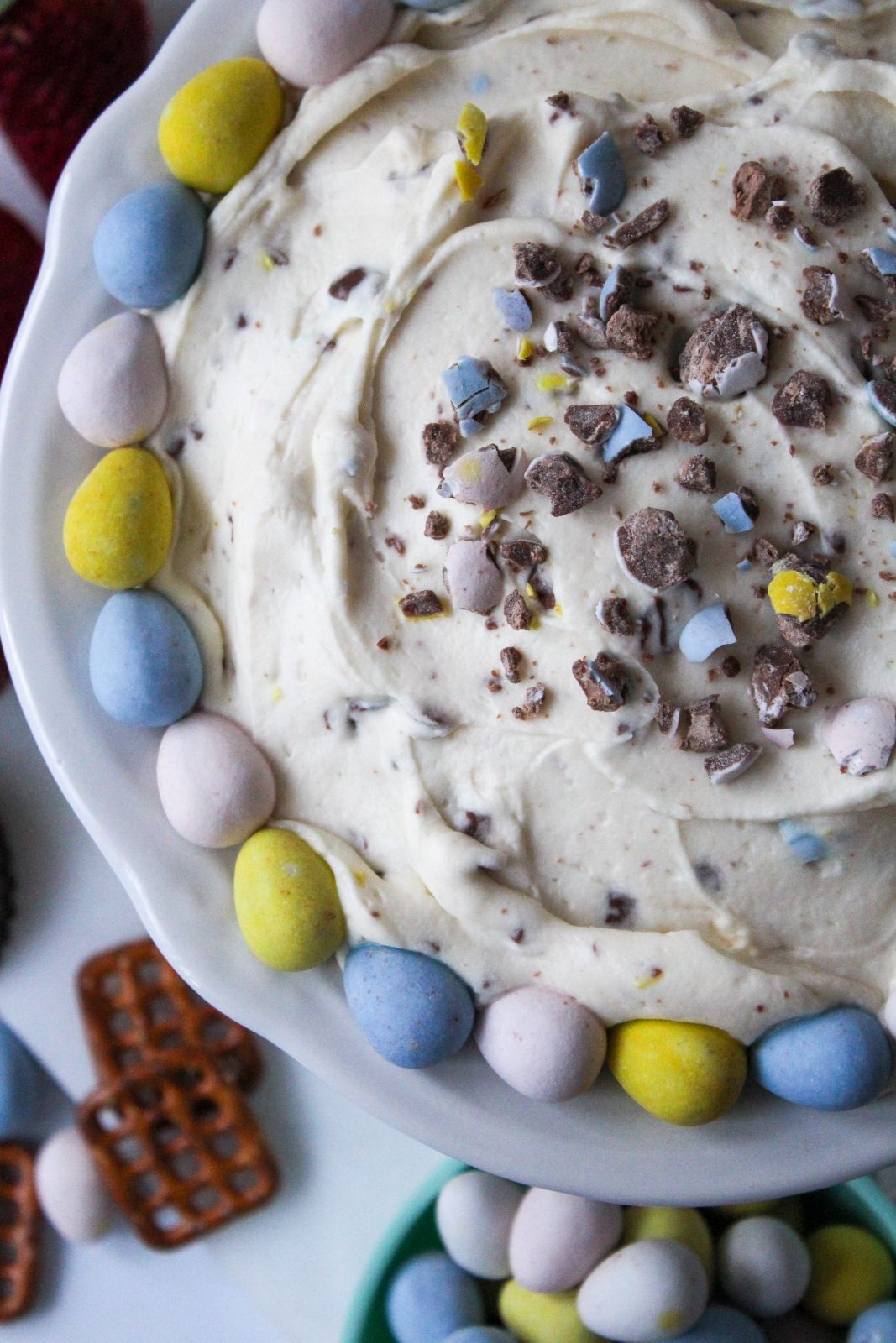 Recipe photo for Cadbury Mini Eggs Whipped Cheesecake Dip showing a creamy colored dip speckled with crushed Cadbury Mini Eggs. Dip is in a scalloped white dish rimmed with whole pastel Cadbury Mini Eggs. In the background are dippers, strawberries, chocolate cookies and pretzels.