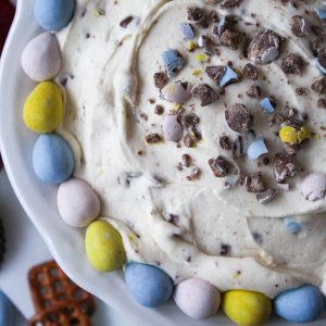 Recipe photo for Cadbury Mini Eggs Whipped Cheesecake Dip showing a creamy colored dip speckled with crushed Cadbury Mini Eggs. Dip is in a scalloped white dish rimmed with whole pastel Cadbury Mini Eggs. In the background are dippers, strawberries, chocolate cookies and pretzels.