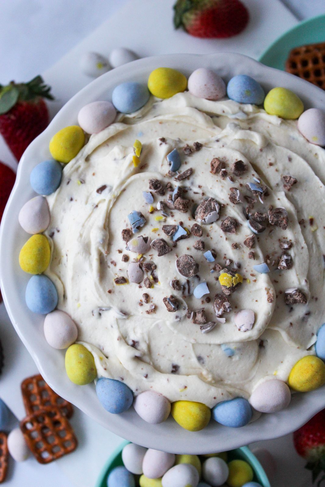 Recipe Image of Cadbury Mini Eggs Whipped Cheesecake Dip. Overhead image of creamy swirled dip in scalloped white bowl. The dip is studded with s sprinkled with crushed Cadbury Mini Eggs candy. Whole pastel colored Cadbury Mini eggs ring around the edge of the bowl in a blue, yellow, pink pattern. In the background are strawberries, pretzels and an aqua cup filled with pastel Cadbury Mini Egg candies.