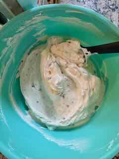 Recipe Image for Cadbury Mini Egg Whipped Cheesecake Dip. A creamy mixture in an ice blue plastic mixing bowl with a black spatula. The creamy whipped white mixture is speckled with crushed pastel Cadbury Mini Eggs.