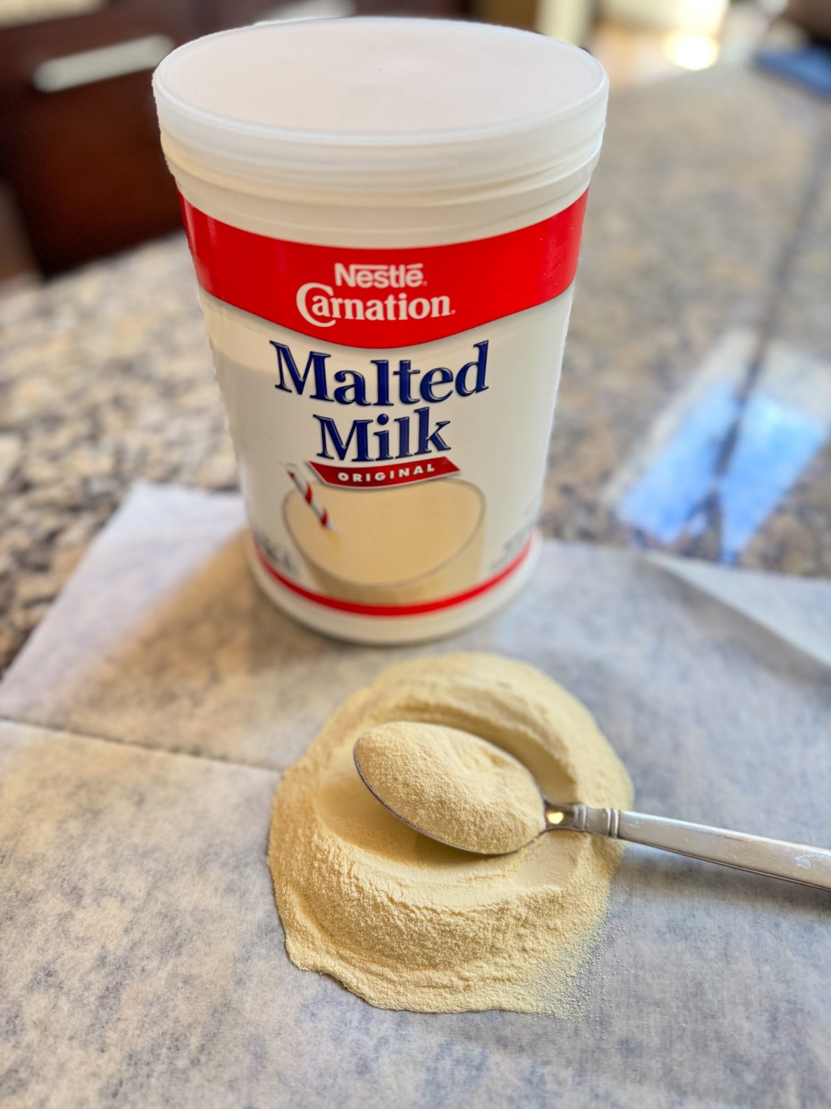 Image showing plastic tub of Carnation Malted Milk Powder sitting on a granite countertop. A sheet of wax paper has a pile of the malted milk powder with a spoon holding a portion.