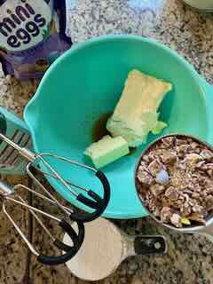 Image showing ingredients for Cadbury Mini Eggs Whipped Cream Cheese Dip Recipe. A aqua sky plastic mixing bowl has cream cheese, half a stick of butter and some vanilla extract. In the back ground is a 1 cup measuring cup with powdered sugar and a 1/2 cup measuring cup with crushed Cadbury Mini Eggs. You can see the flex edge beaters for the KitchenAid Hand Mixer.