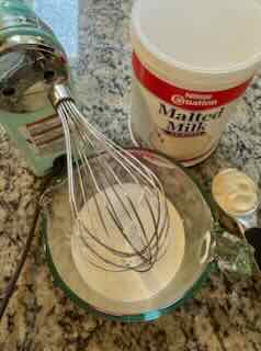 Image showing ingredients for making Malted Whipped Cream. with an ice blue KitchenAid 7-speed hand mixer fitted with a Pro-Whisk. A container for Carnation Malted Milk powder and a measuring cup with malted milk powder. In a mixing bowl is heavy cream.