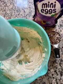 Recipe Image for Cadbury Mini Egg Whipped Cheesecake Dip. An ice blue KitchenAid Hand Mixer is mixing a creamy mixer in a matching blue mixing bowl. In the background is a purple bag of Cadbury Mini Eggs.