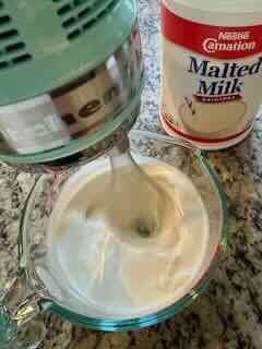 Image showing an ice blue KitchenAid 7-Speed Hand Mixer with Pro Whisk Whipping Cream. A can of Carnation Malted milk powder is in the background.