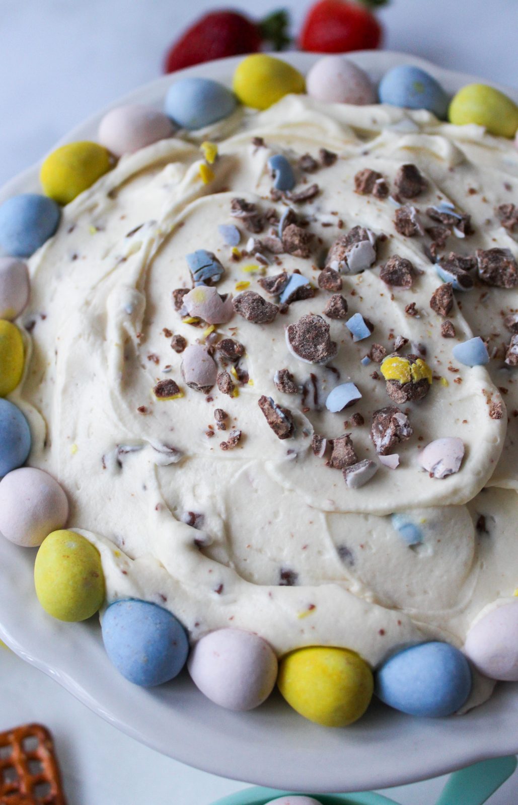 Recipe Image of Cadbury Mini Eggs Whipped Cheesecake Dip. Close up image of creamy swirled dip in scalloped white bowl. The dip is studded with s sprinkled with crushed Cadbury Mini Eggs candy. Whole pastel colored Cadbury Mini eggs ring around the edge of the bowl in a blue, yellow, pink pattern. In the background are strawberries and pretzels.