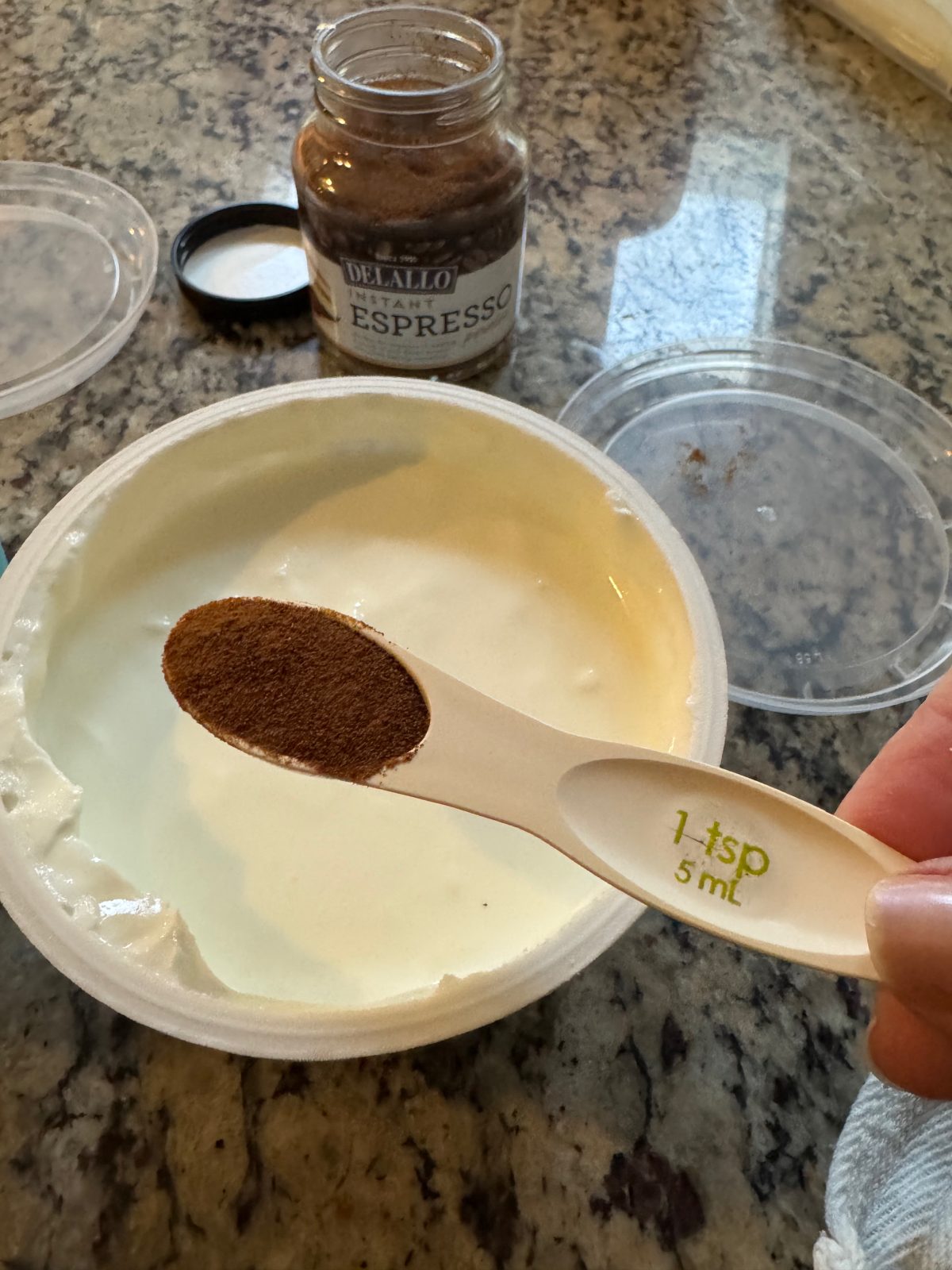 recipe image for Viral Greek Yogurt Biscoff Cheesecake (tiramisu version) Shows top view of yogurt tub with spoonful of espresso powder about to be added to it.