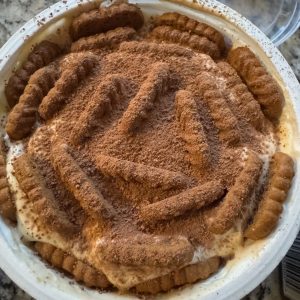 recipe image for Viral Greek Yogurt Biscoff Cheesecake (tiramisu version) Shows top view of yogurt tub with biscoff cookies inserted into it and sprinkled with cocoa powder