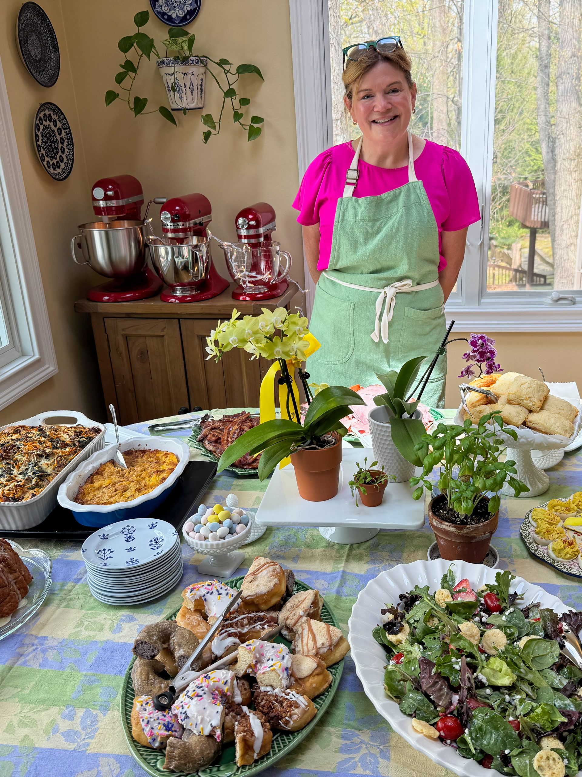 Image shows Tina Verrelli of epicuricloud.com standing behind her Easter brunch buffet table. The table has a pastel checkered table cloth with platters showing salad, sweet pastries, bacon, ham, deviled eggs and a floral and herb arrangement as the center piece. In the background are 3 sizes of KitchenAid Stand Mixers on a wooden cabinet.
