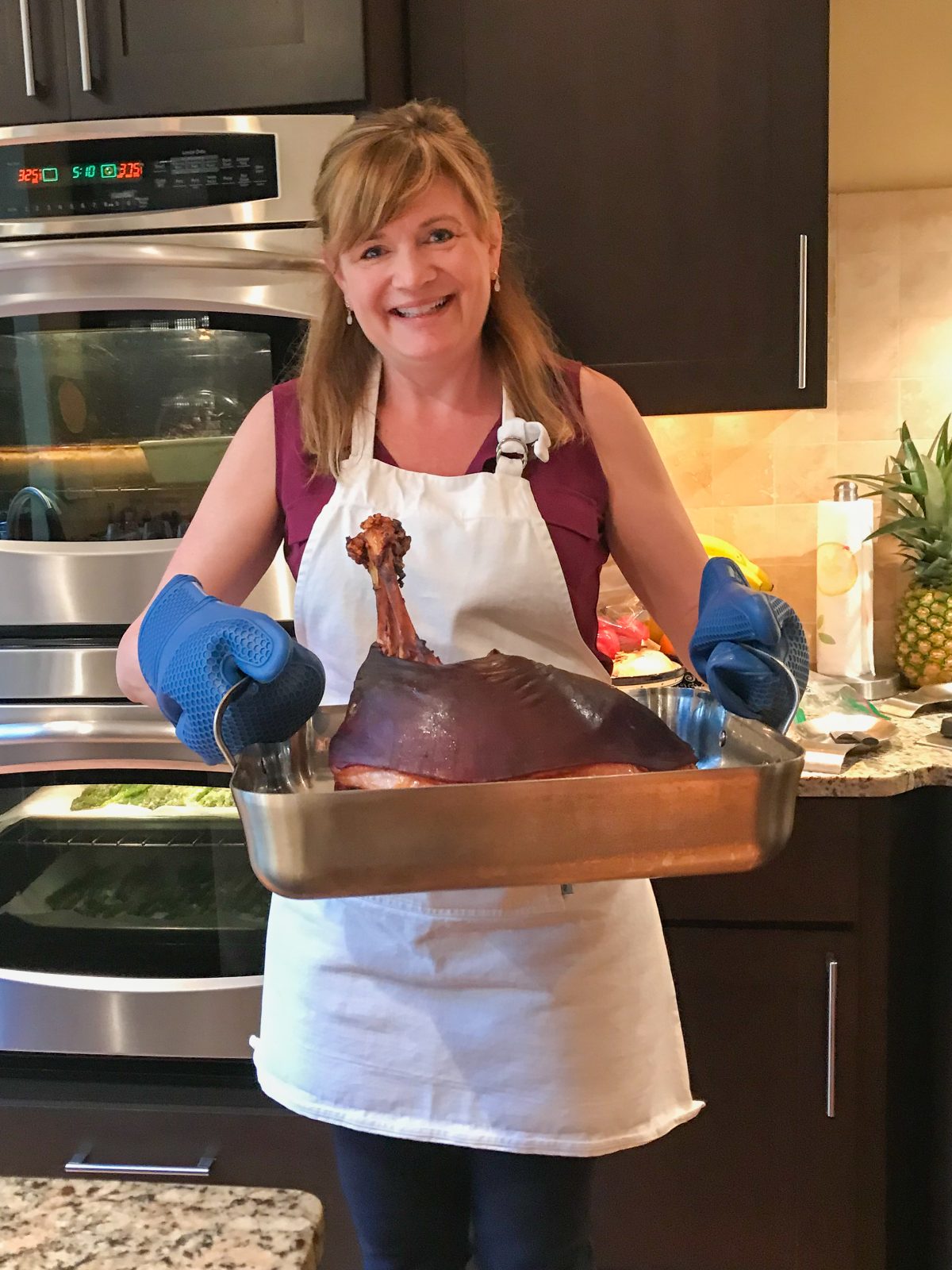 Tina Verrelli of epicuricloud.com holding a large baked bone in ham with skin.  The ham is a large stainless steel roasting pan.  Tina is wearing a sleeveless purple top with a white full apron.  She has large blue silicone oven mitts protecting her hands.  In the background you can see her kitchen with double ovens and espresso colored cabinetry.