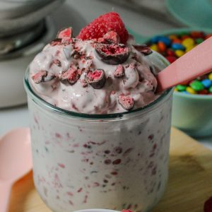 Recipe Photo in close up and vertical orientation for How to Make a Blended Blizzard Style Ice Cream Treat with Your Stand Mixer showing close up shot of KitchenAid 5qt stand mixer shiny stainless steel bowl. A short glass is filled with the pale pink blizzard mixture. There is a pink plastic spoon in the glass. Fresh raspberries and TruFru (chocolate covered freeze dried berries) candy garnish the treat. There are bowls of candies and cookies in the background.