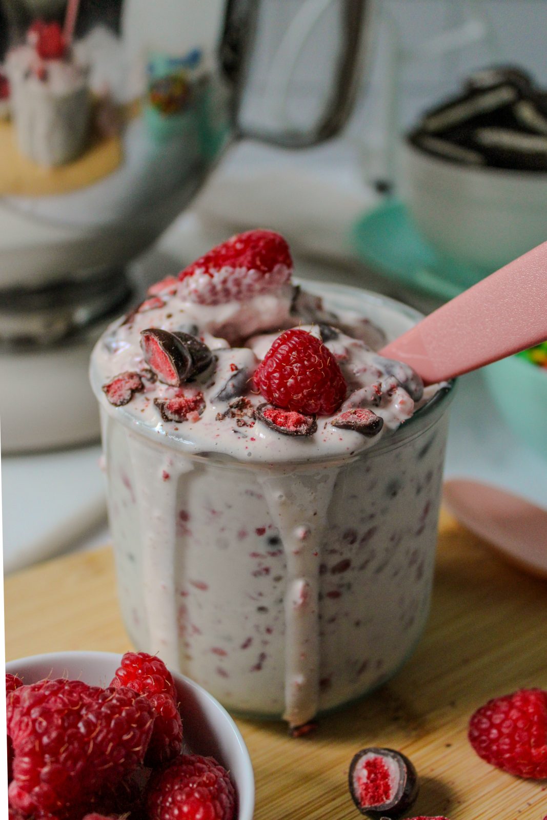 Recipe Photo for How to Make a Blended Blizzard Style Ice Cream Treat with Your Stand Mixer showing close up shot of KitchenAid 5qt stand mixer shiny stainless steel bowl. A short glass is filled with the pale pink blizzard mixture. There is a pink plastic spoon in the glass and drips of the creamy mixture are overflowing the glass. Fresh raspberries and TruFru (chocolate covered freeze dried berries) candy garnish the treat. There are bowls of candies and cookies in the background.
