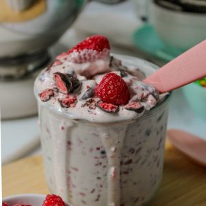 Recipe Photo for How to Make a Blended Blizzard Style Ice Cream Treat with Your Stand Mixer showing close up shot of KitchenAid 5qt stand mixer shiny stainless steel bowl. A short glass is filled with the pale pink blizzard mixture. There is a pink plastic spoon in the glass and drips of the creamy mixture are overflowing the glass. Fresh raspberries and TruFru (chocolate covered freeze dried berries) candy garnish the treat. There are bowls of candies and cookies in the background.