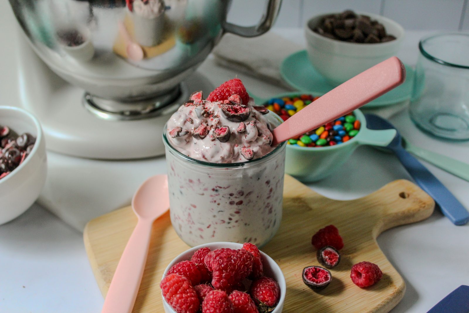Recipe Photo in horizontal orientation for How to Make a Blended Blizzard Style Ice Cream Treat with Your Stand Mixer showing close up shot of KitchenAid 5qt stand mixer shiny stainless steel bowl. A short glass is filled with the pale pink blizzard mixture. There is a pink plastic spoon in the glass. Fresh raspberries and Tru Fru (chocolate covered freeze dried berries) candy garnish the treat. There are bowls of candies and cookies in the background.