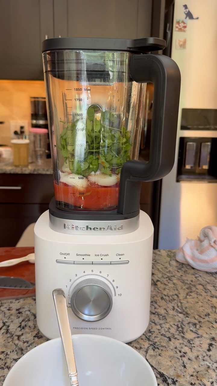 Recipe Photo for How to Make Salsa with Canned Tomatoes. The blender is porcelain white. the glass blender jar is layered with tomatoes, onions, jalapeño, cilantro and spices. The blender is sitting on a neutral tan granite countertop with a stainless steel refrigerator in the background.