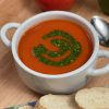 Recipe photo for Quick and Easy Tomato Soup. Vertical image showing white ceramic bowl with handles containing red smooth tomato soup with a swirl of green pesto. The bowl is set on a wooden cutting board and surrounded with slices of baguette on a blue and green cloth napkin, a shiny silver spoon and a ripe fresh tomato.