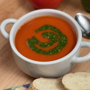 Recipe photo for Quick and Easy Tomato Soup. Vertical image showing white ceramic bowl with handles containing red smooth tomato soup with a swirl of green pesto. The bowl is set on a wooden cutting board and surrounded with slices of baguette on a blue and green cloth napkin, a shiny silver spoon and a ripe fresh tomato.