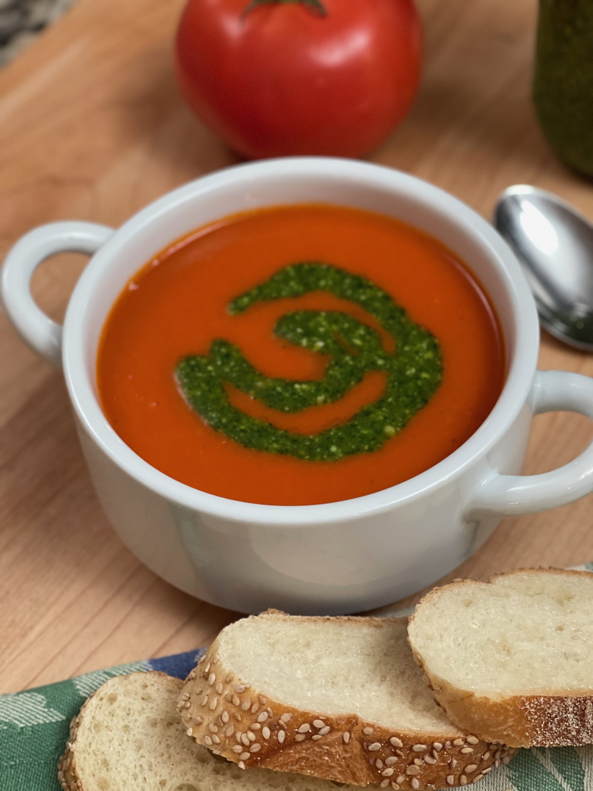 Recipe photo for Quick and Easy Tomato Soup. Vertical image showing white ceramic bowl with handles containing red smooth tomato soup with a swirl of green pesto. The bowl is set on a wooden cutting board and surrounded with slices of baguette on a blue and green cloth napkin, a shiny silver spoon and a ripe fresh tomato.