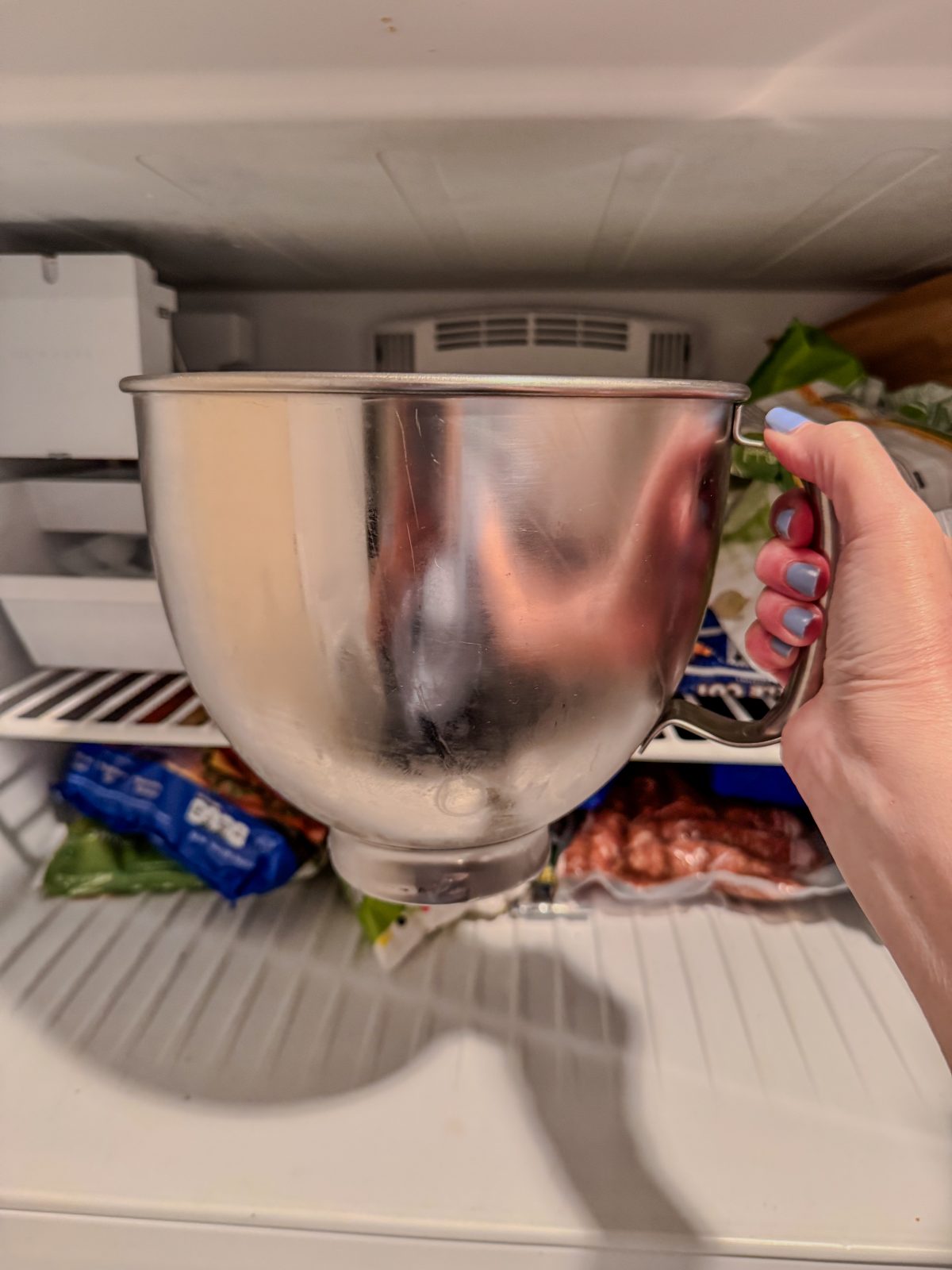 Recipe Process Photo for How to Make a Blended Blizzard Style Ice Cream Treat with Your Stand Mixer showing taking stainless steel KitchenAid 5qt stand mixer bowl from the freezer. Typical top freezer with 1 shelf, ice maker and bags of frozen vegetables.
