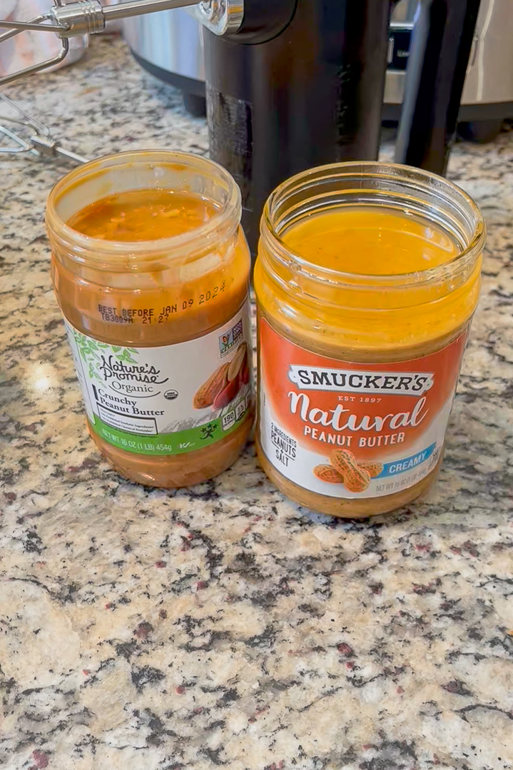 Two jars of natural peanut butter (Smuckers and Nature's Promise) on black speckled granite countertop. Black KitchenAid cordless hand mixer in the background.