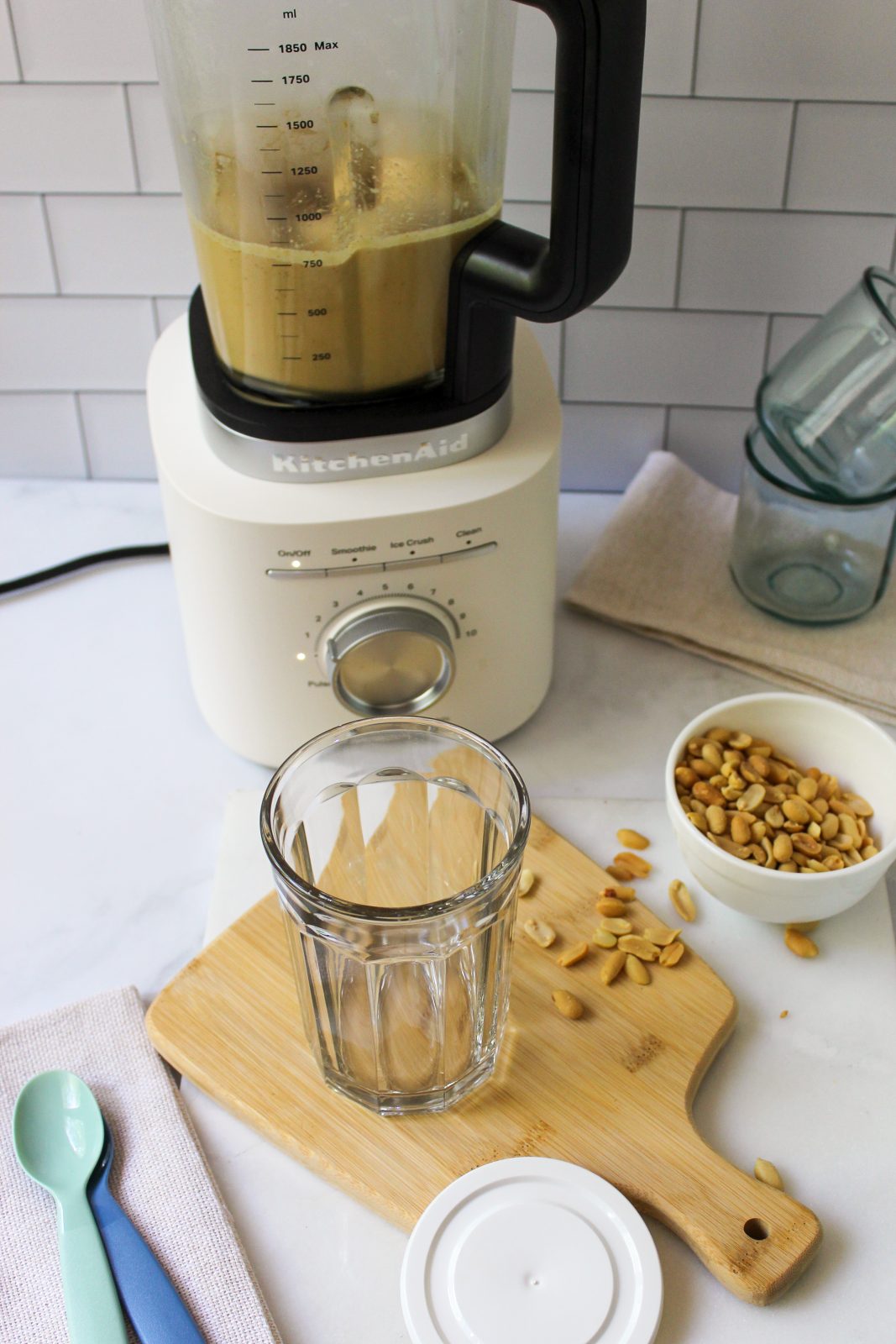 Recipe Photo for Homemade Peanut Butter. Shows peanut butter blended and ready to pour from the Porcelain White KitchenAid Pure Power Blender with Glass Jar. The clear jar the peanut butter will be poured into is set upon a small bamboo cutting board and there is a small white bowl with peanuts in it. The countertop is marble and the backsplash is white subway tile.
