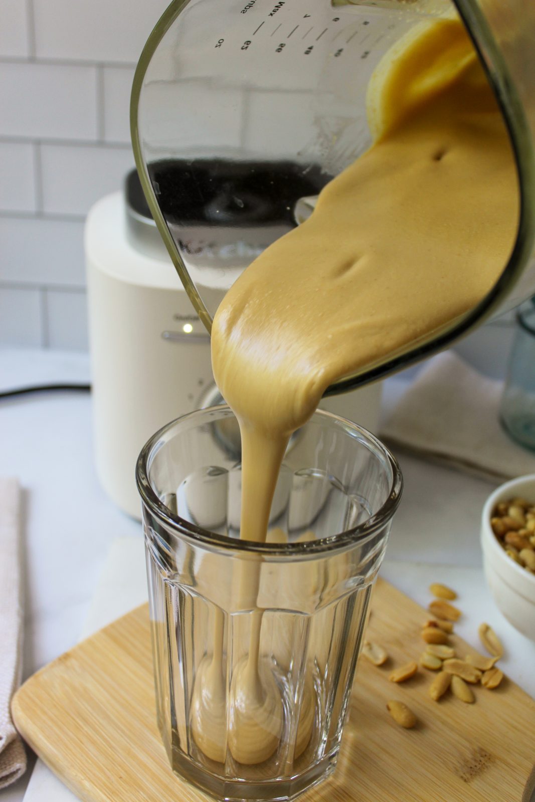 Recipe Photo for Homemade Peanut Butter. Shows starting to pour the peanut butter from the glass KitchenAid Pure Power Blender Jar. You can see the Porcelain White Blender Base in the background. The clear jar the peanut butter is being poured into is set upon a small bamboo cutting board and there is a small white bowl with peanuts in it. The countertop is marble and the backsplash is white subway tile.