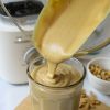 Recipe Photo for Homemade Peanut Butter. Shows pouring the peanut butter from the glass KitchenAid Pure Power Blender Jar. You can see the Porcelain White Blender Base in the background. The clear jar the peanut butter is being poured into is set upon a small bamboo cutting board and there is a small white bowl with peanuts in it. The countertop is marble and the backsplash is white subway tile.