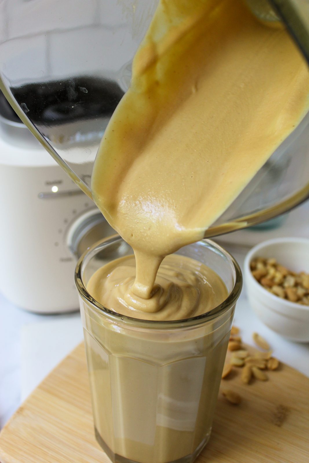 Recipe Photo for Homemade Peanut Butter. Shows pouring the peanut butter from the glass KitchenAid Pure Power Blender Jar. You can see the Porcelain White Blender Base in the background. The clear jar the peanut butter is being poured into is set upon a small bamboo cutting board and there is a small white bowl with peanuts in it. The countertop is marble and the backsplash is white subway tile.