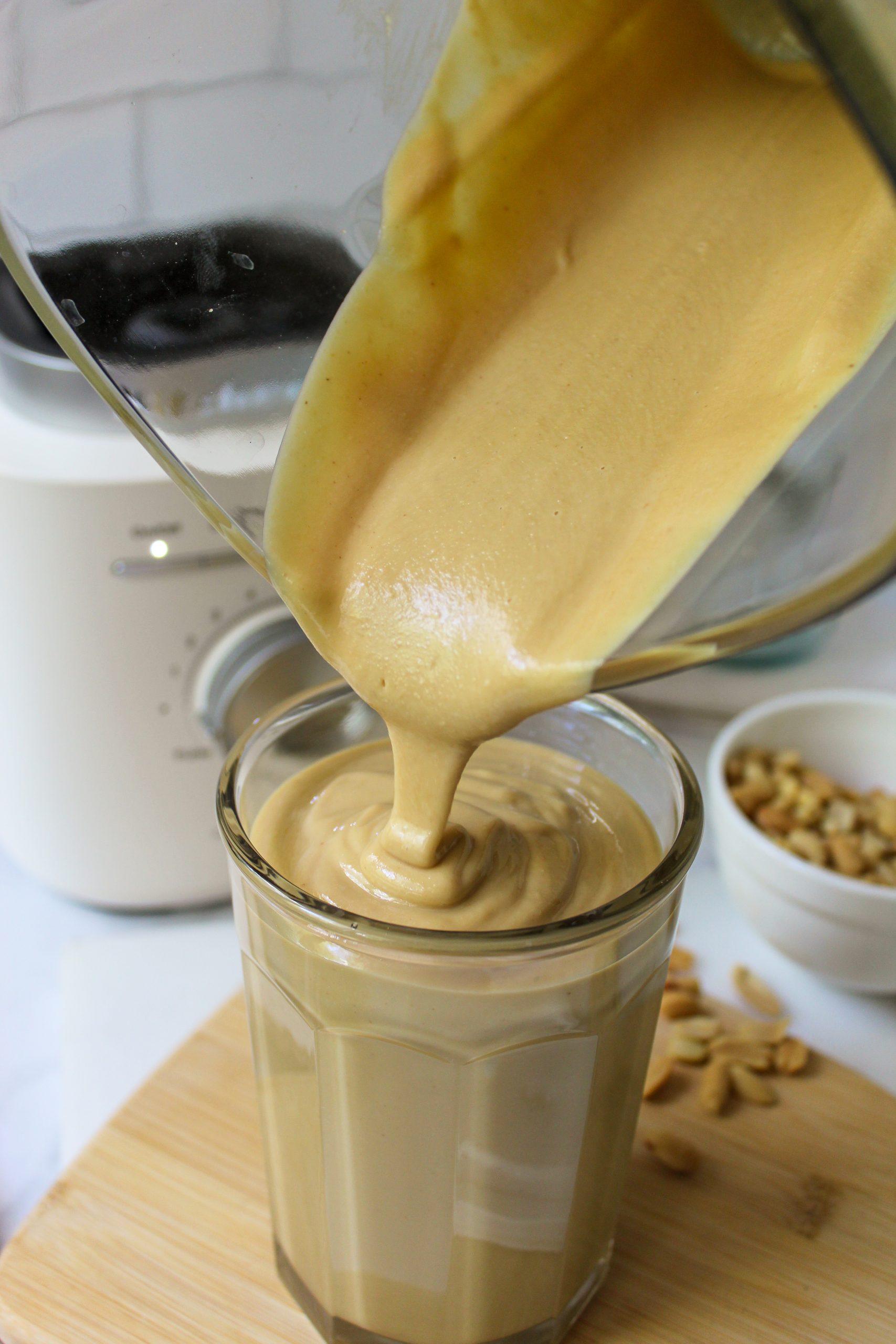 Recipe Photo for Homemade Peanut Butter. Shows pouring the peanut butter from the glass KitchenAid Pure Power Blender Jar. You can see the Porcelain White Blender Base in the background. The clear jar the peanut butter is being poured into is set upon a small bamboo cutting board and there is a small white bowl with peanuts in it. The countertop is marble and the backsplash is white subway tile.