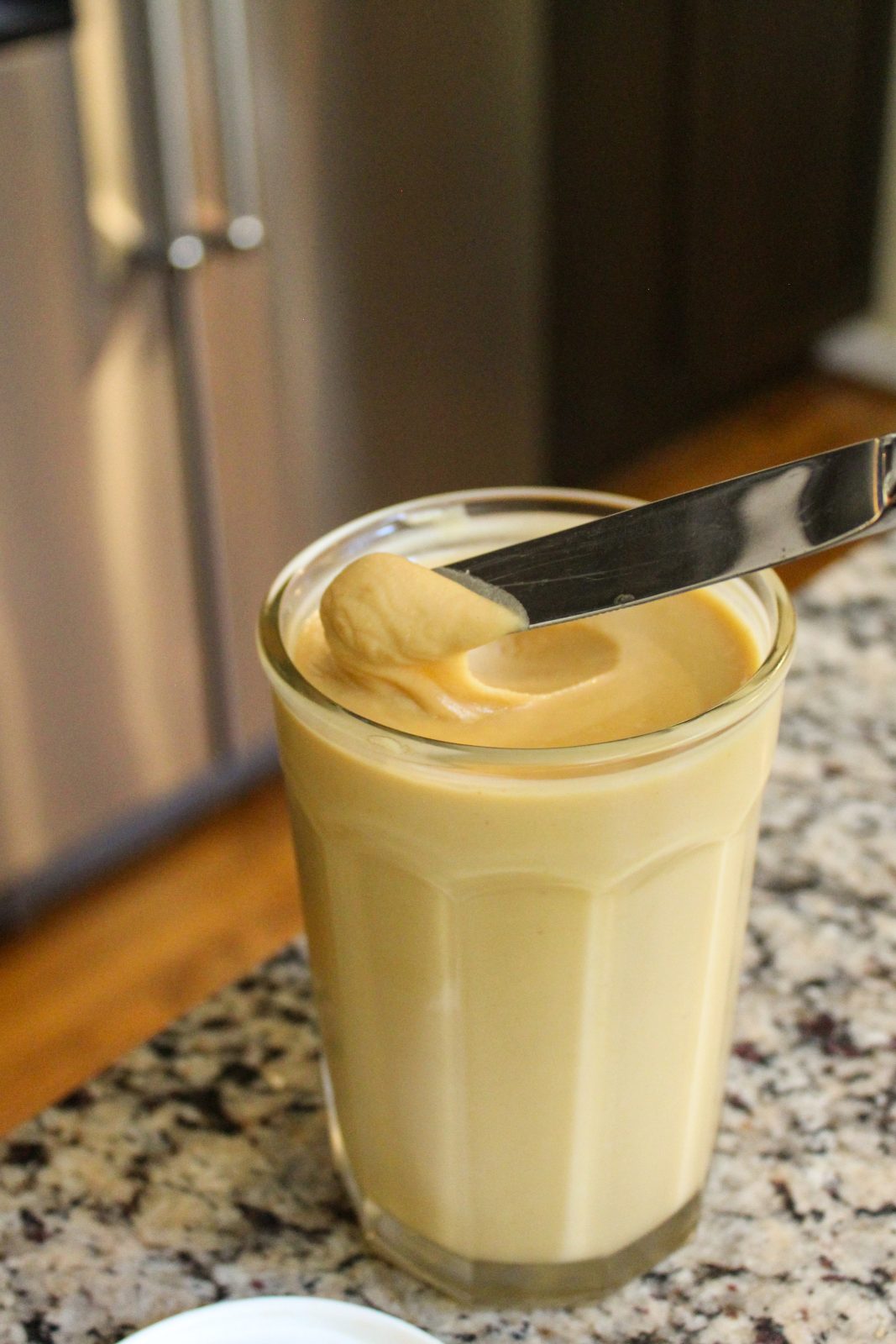 Recipe Photo for Homemade Peanut Butter. Shows glass jar of homemade peanut butter with a knife dipping into the jar to show spreadability after being chilled in refrigerator for 24 hours. The jar is on a speckled beige and black granite countertop, in the background you can see a stainless steel side by side refrigerator and hard wood flooring.