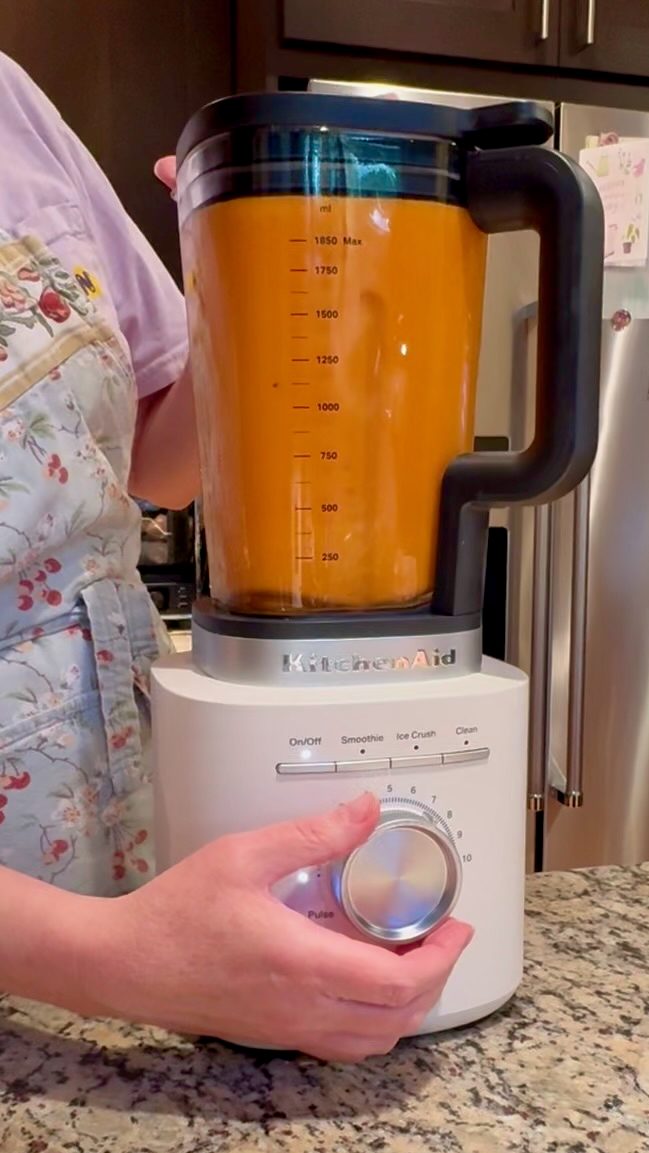 Recipe photo for Quick and Easy Tomato Soup. Vertical photo showing a Porcelain White KitchenAid Pure Power Blender blending orange colored creamy Quick and Easy Tomato Soup. I am standing behind the blender in a floral apron, lavender t-shirt and my hand is on the blender knob. You can see a speckled granite countertop under the blender.