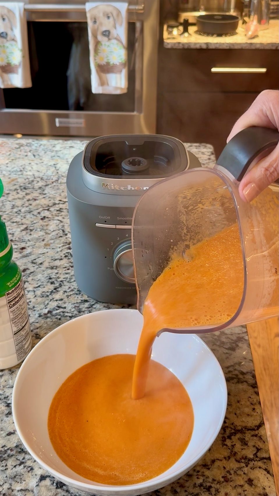 Recipe photo for Quick and Easy Tomato Soup. Vertical photo showing a gray KitchenAid Pure Power Blender pouring blended orange colored creamy Quick and Easy Tomato Soup into a white bowl. You can see a speckled granite countertop under the blender.